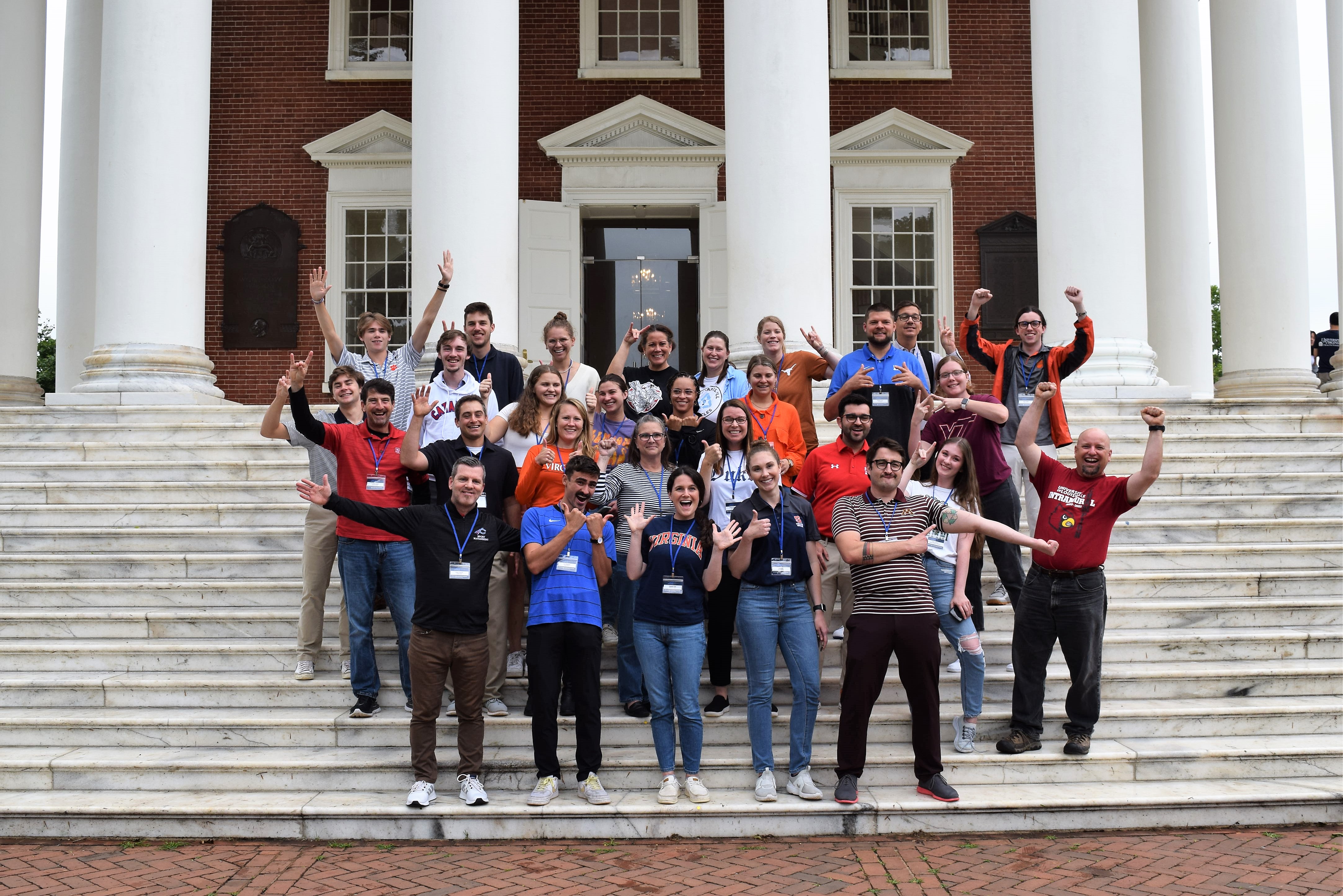 29 smiling people standing as a group on the steps of the Rotunda. While holding out their arms or making hand symbols representing their Universities, they all wear university or association branded shirts.