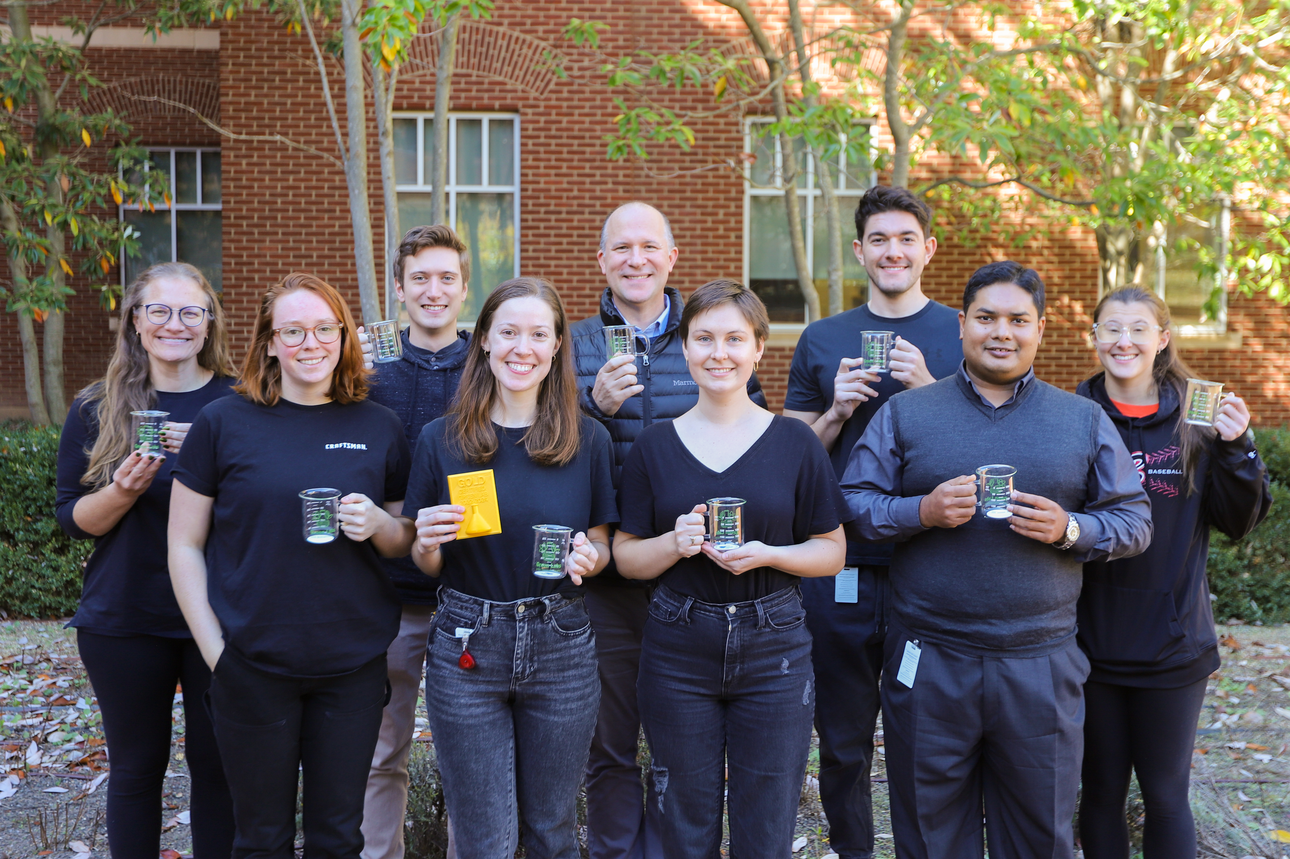 A group of nine researchers stands together smiling and holding their Green Labs beaker mug and plaques. 