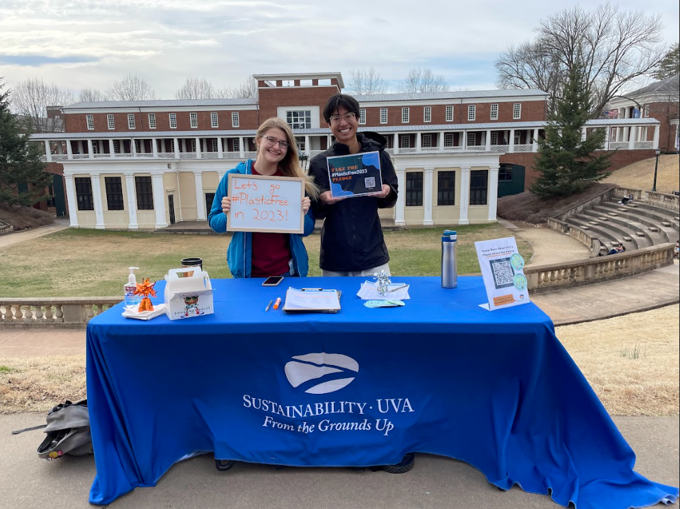Two students tabling for #PlasticFree2023 campaign at UVA's Amphitheater. One has long blonde hair and fair skin and hold a whiteboard that says 'Let's Go Plastic Free In 2023'. The other has short dark hair with olive skin and is holding a piece of paper that says #PlasticFree2023 with a QR code to sign the pledge. They are both wearing coats and stand behind a blue tablecloth.