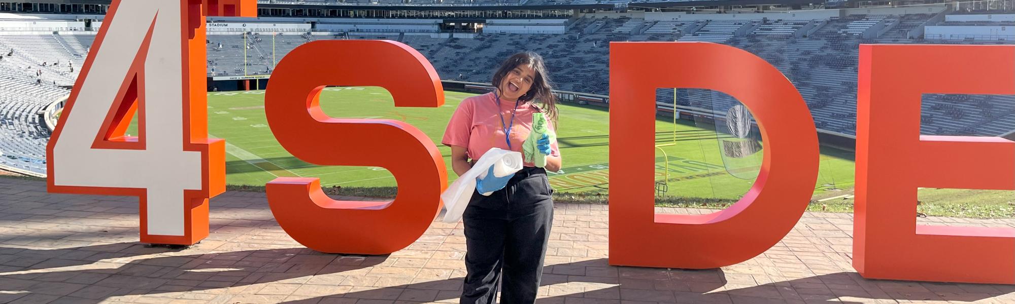 Student volunteer smiles with compost/recycling bags in front of a sign that says 4 SIDE and Scott Stadium