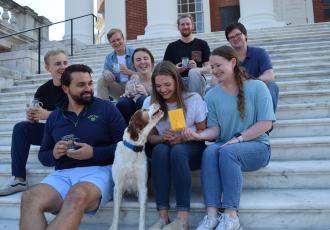 A group of 8 researchers are sitting on the steps of the Rotunda with a cute dog with long, soft, fluffy ears. They're all smiling and holding their Certification plaque and beaker mugs.