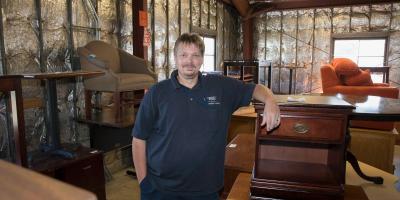 An image of Glenn from the ReUse Store. He is smiling at the camera while leaning on a wooden dresser. The background shows multiple furniture items and walls that have silver insulation.