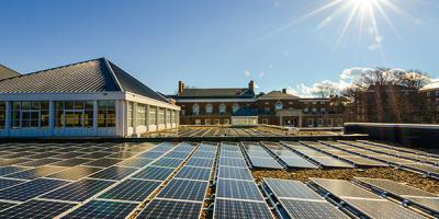Image of rectangular solar panels lining a building roof at UVA. The background shoes the surrounding red brick buildings and a clear blue sky.