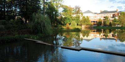 An image of the Dell Pond at UVA shows dark blue water surrounded by tall green trees that are casting shadows on the water surface. A red brick UVA building is in the background with a cloudy blue sky.