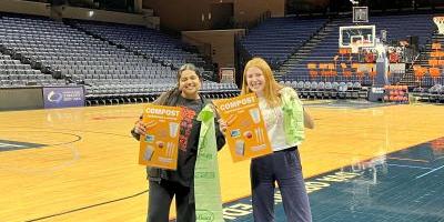 Two student employees display compost bags and signs on the court of John Paul Jones Arena